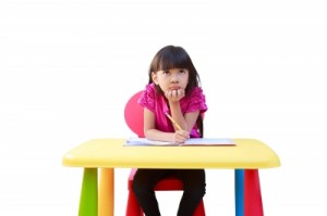 Young girl sitting at plastic desk, thinking and writing in a notebook