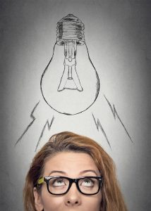 Headshot thinking businesswoman with glasses having an idea looking up with light bulb over her head isolated grey wall background. Human face expressions, emotions. Creativity concept
